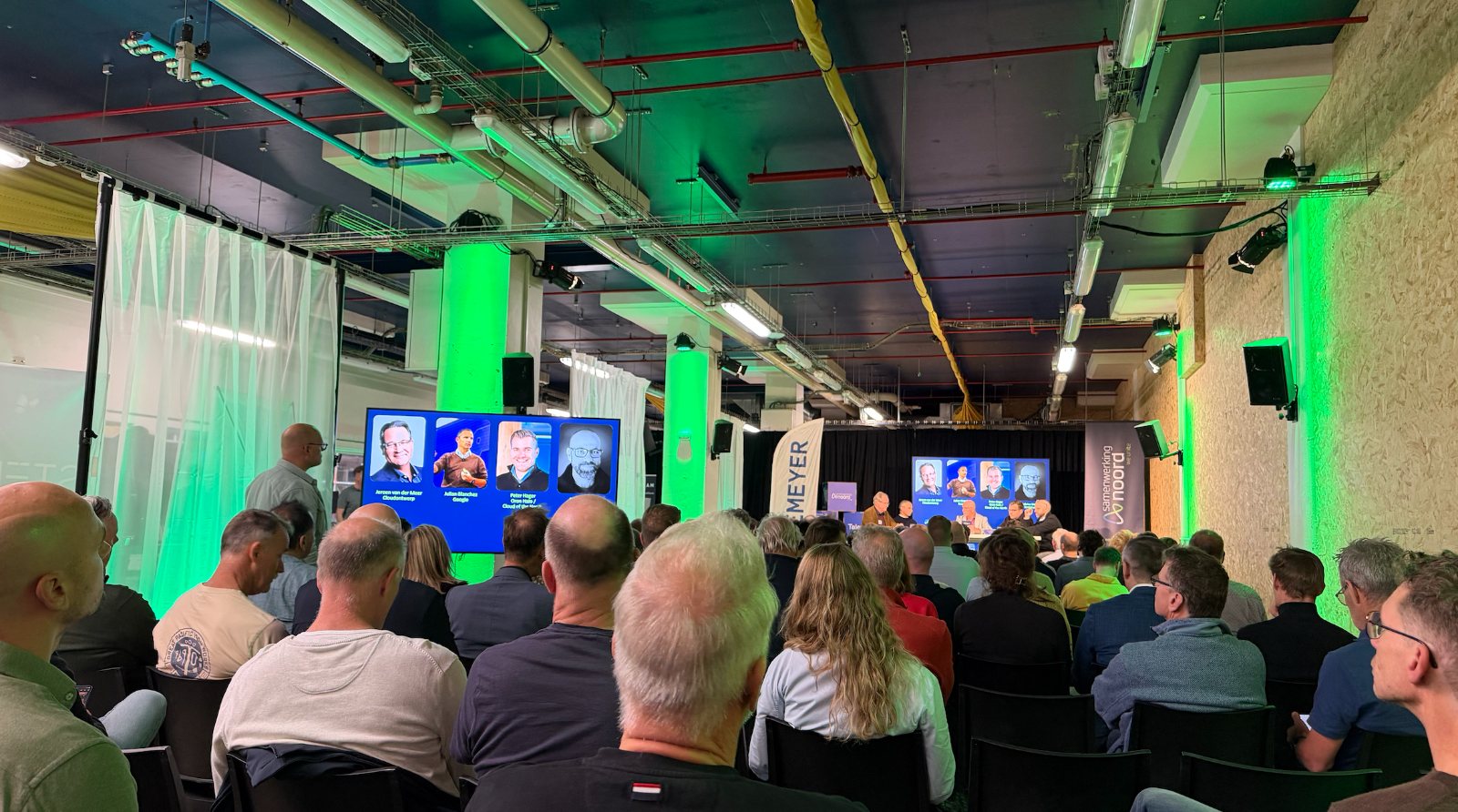 Volle zaal in de Niemeyer fabriek, groen aangelicht, kijkend naar de talkshow met op het scherm de vier gasten.